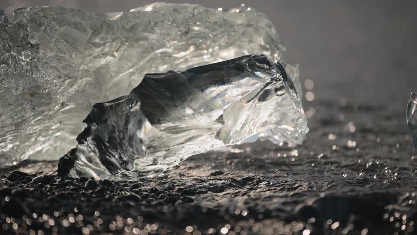 A close view of a transparent ice block resting on a black pebble beach with strong sunlight refracting through the textured frozen surface