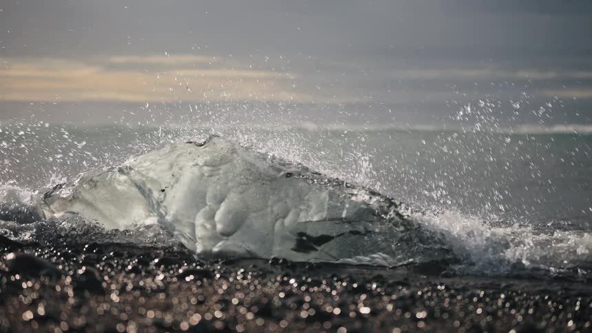 Transparent ice chunk struck by waves on dark beach at dusk
