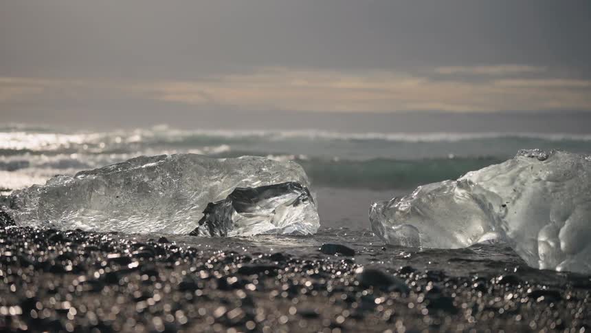 Translucent ice chunks on pebble beach with calm sea and cloudy sky in the background