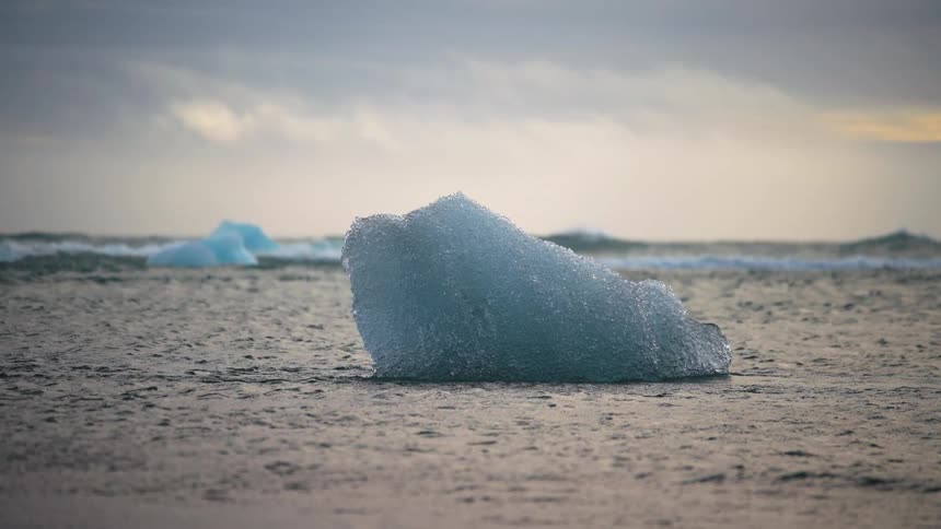 Blue ice chunk on dark sand with ocean in the background under cloudy sky