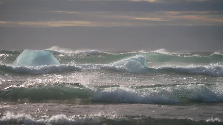 Large turquoise waves crest and crash around two blue icebergs floating in a stormy cold ocean beneath a heavily clouded sky