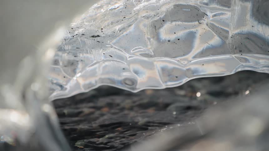 A close view of dark rocks under shallow clear water seen through an opening in textured ice showing colorful light refractions