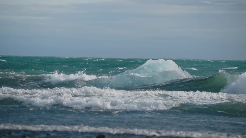 Large turquoise waves crest and crash around two blue icebergs floating in a stormy cold ocean beneath a heavily clouded sky