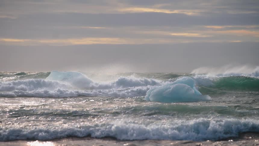 Large turquoise waves crest and crash around two blue icebergs floating in a stormy cold ocean beneath a heavily clouded sky