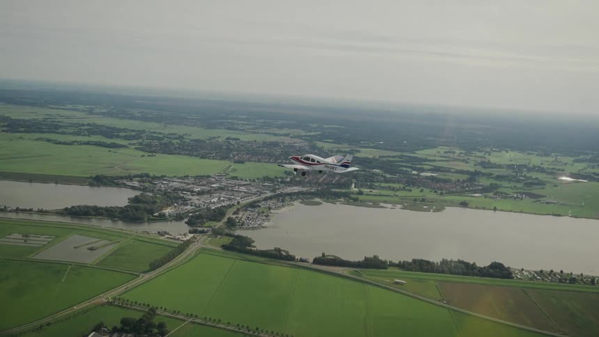 A wide aerial view shows a propeller plane flying low over a town sitting between two lakes and surrounded by lush green rural fields