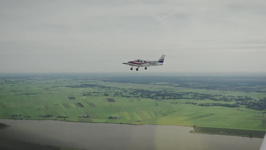 An aerial view of a single engine plane flying over vast flat green farmlands bordered by a wide river under a light cloudy sky