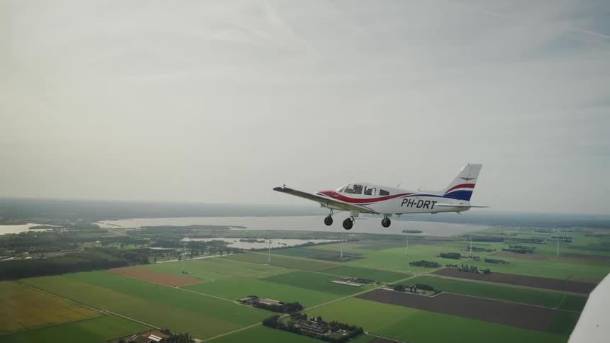 A white red and blue propeller plane flies with its landing gear down over geometric green fields a lake and wind turbines