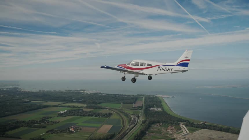 A small propeller airplane flies with its landing gear down over green fields and a coastal embankment separating the land from the ocean