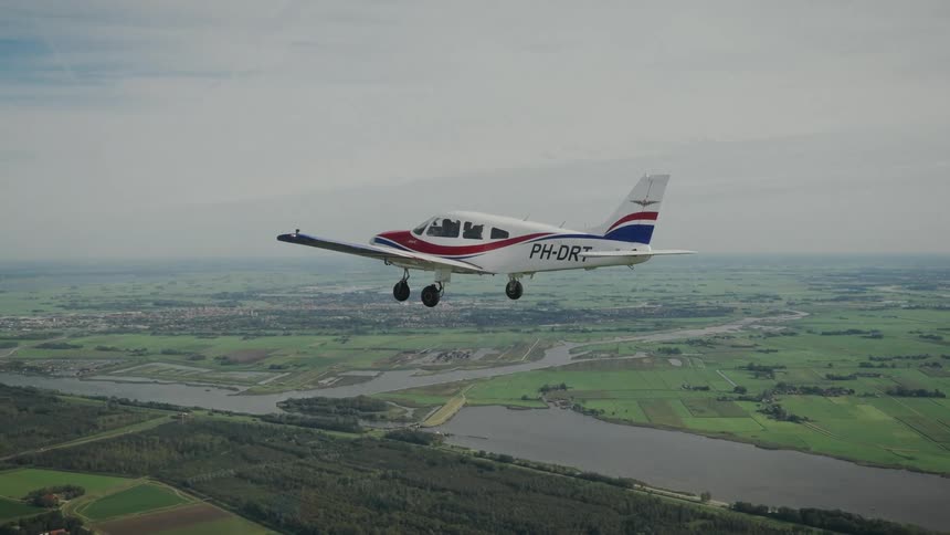 A white red and blue single engine plane flies low with its landing gear down over a winding river and lush green agricultural fields