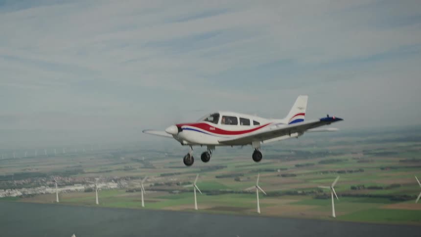 A small airplane flies above fields lined with wind turbines