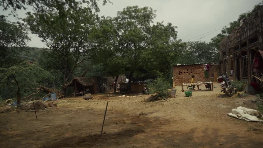 A wide view of a rustic rural dwelling with a dirt yard a motorcycle and a cot near a brick building surrounded by dense green trees and foliage