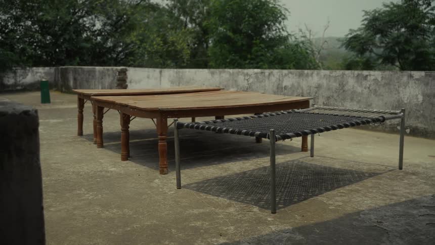 Two tables with decorative carved legs and a cot with a simple metal frame with black woven strapping resting on a bare sunlit rooftop with a concrete parapet wall and dense green foliage visible in the background under an overcast sky
