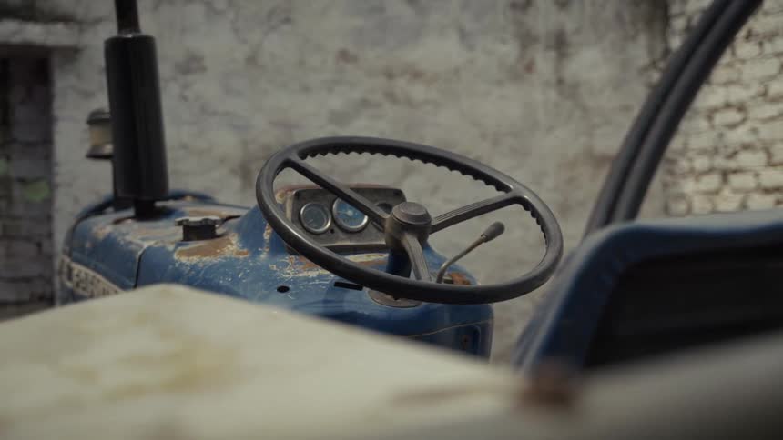 A black steering wheel and dashboard of an old faded blue tractor that is rusting and parked in front of a rough pale brick wall