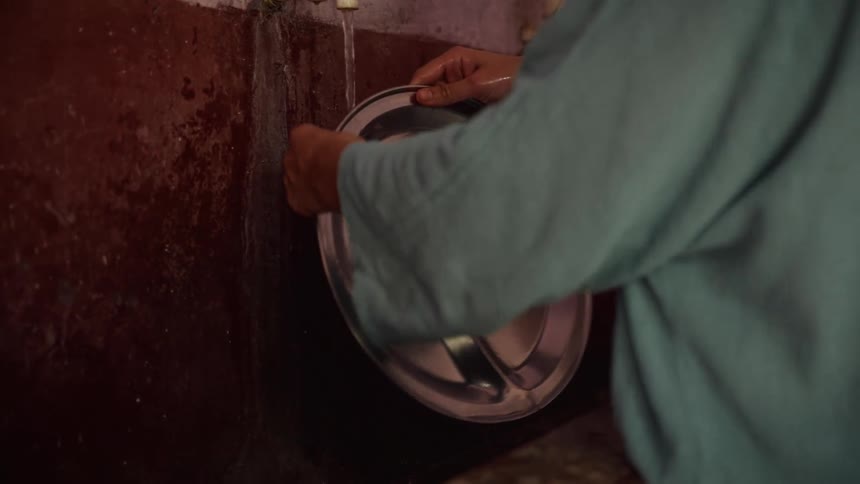 A woman wearing a green dress washes a stainless steel plate at an outdoor faucet beside a wall at midday