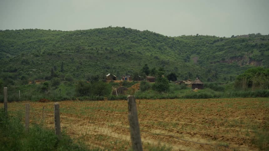 Plowed land fills the foreground with small huts at the base of green hills