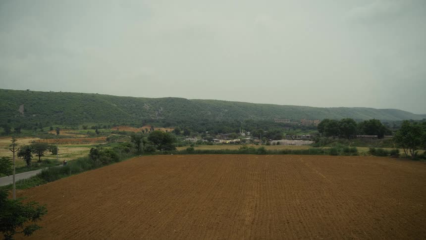 Plowed field fills the foreground with a village at the base of green hills under an overcast sky