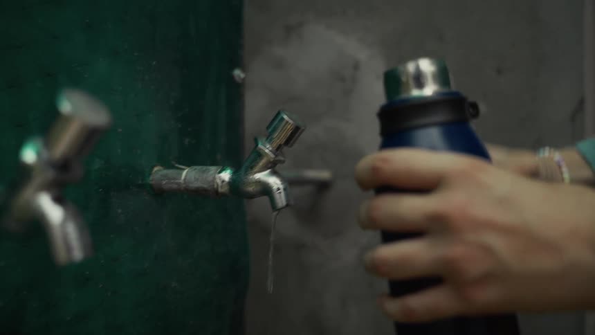 A person filling a water bottle at a metal tap and then capping the bottle