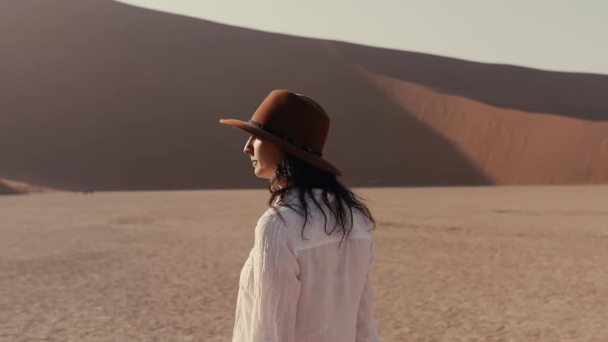A woman in a hat slowly walks across a desert landscape