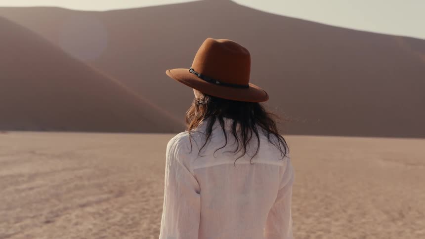 A woman viewed from behind and wearing a widebrimmed brown hat and a white shirt walks slowly across the flat sandy ground towards the distant massive sand dunes of a desert landscape