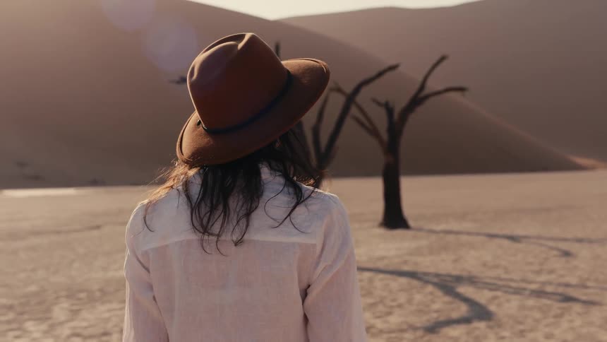 Woman wearing a shite shirt and a brown hat walking on a plain area of land with mountains on the background outdoors during day time