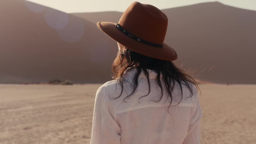 A woman viewed from behind and wearing a widebrimmed brown hat and a white shirt walks slowly across the flat sandy ground towards the distant massive sand dunes of a desert landscape