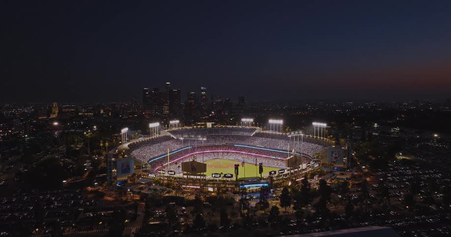 The video is an aerial wide shot of a brightly lit baseball stadium likely dodger stadium hosting a night game the stadium is surrounded by dark parking lots and trees with the illuminated skyline of a city visible in the background