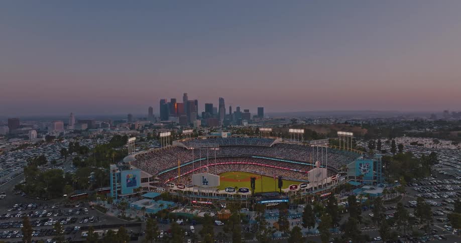 Aerial view of dodger stadium glows under bright lights as dusk settles over the city with the downtown skyline rising majestically in the background