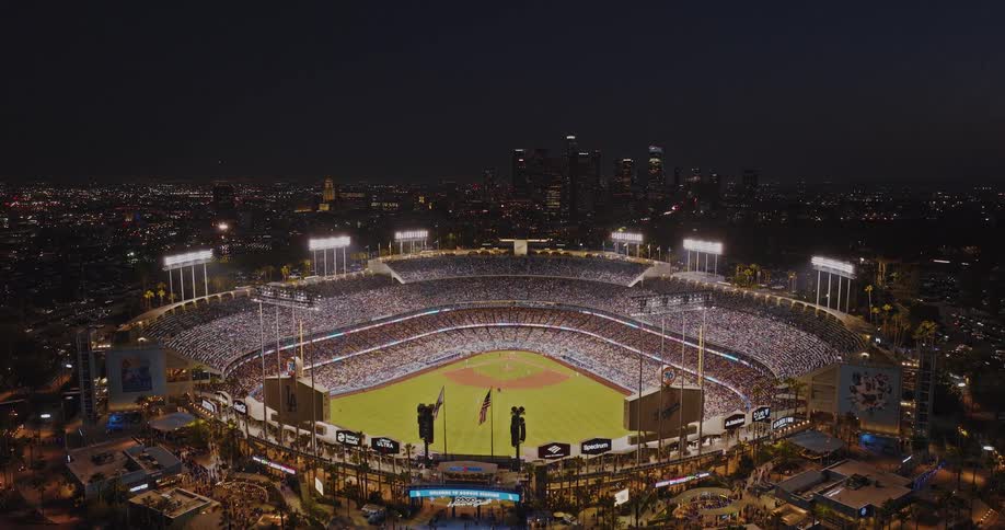 A highangle aerial shot captures a brightly illuminated stadium during a night game packed with a large crowd set against the dark backdrop of the surrounding city lights and the city skyline