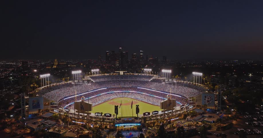 An aerial view of a packed baseball stadium glowing at night with a city skyline behind it