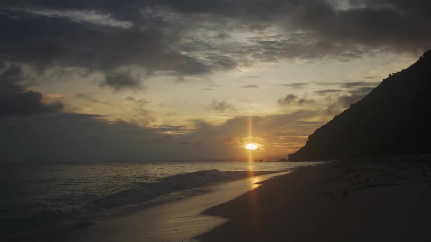 Sun shines from a red horizon casting the beachside hill into silhouette under a cloudy sky