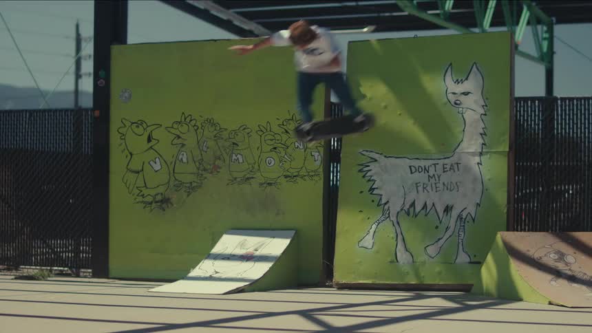 A skateboarder in a white shirt and dark pants launches off a small kicker ramp midair and twisting past a tall green wall painted with a cartoon llama at an outdoor skatepark