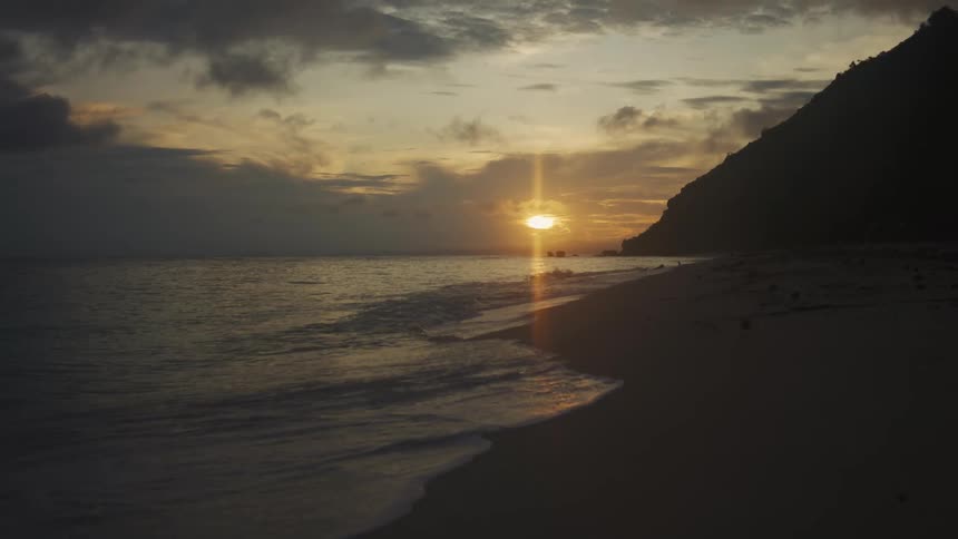A sunset or sunrise over a beach where the golden sun is low on the horizon casting a bright flare over the ocean waves and reflecting on the wet sand a dark steep hill or mountain dominates the right side of the frame contrasting with the light sky