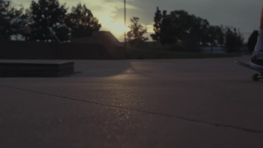 A skateboarders feet push off and glide across the smooth concrete of a skatepark silhouetted by the warm glow of the setting sun