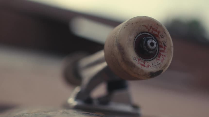 An extremely tight lowangle closeup shot focuses on a dirty white skateboard wheel and its silver truck assembly with the rest of the skateboard deck blurring into the background