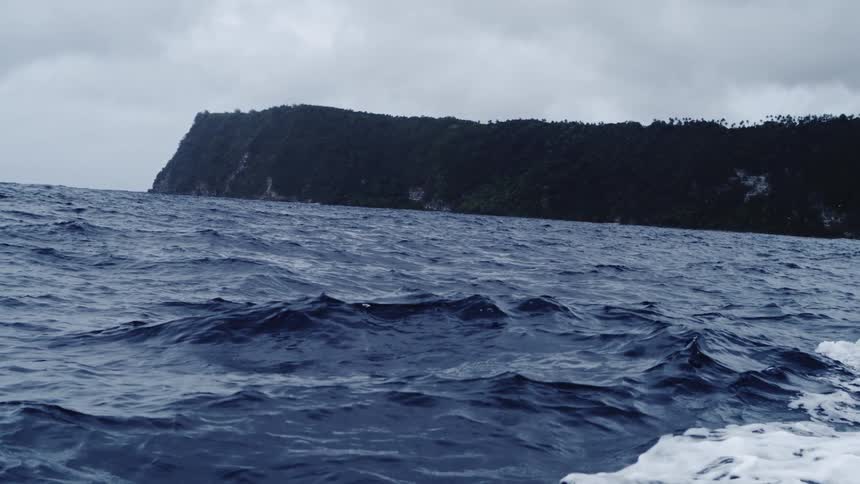 The surface of the blue water of an ocean with small ripples moving and a large stretch of hill in the background