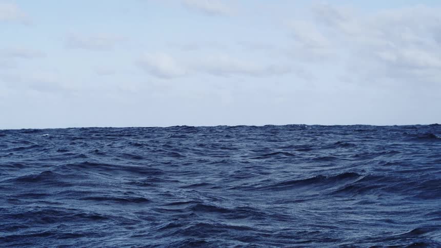 A vast expanse of deep dark blue ocean with choppy waves filling the lower twothirds of the frame meeting a clear horizon beneath a light blue sky scattered with thin white clouds