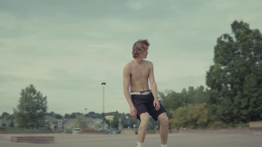 A shirtless male skateboarder successfully executes a jump down a large set of concrete stairs landing on his board and rolling away onto the flat ground of the skatepark under an overcast sky