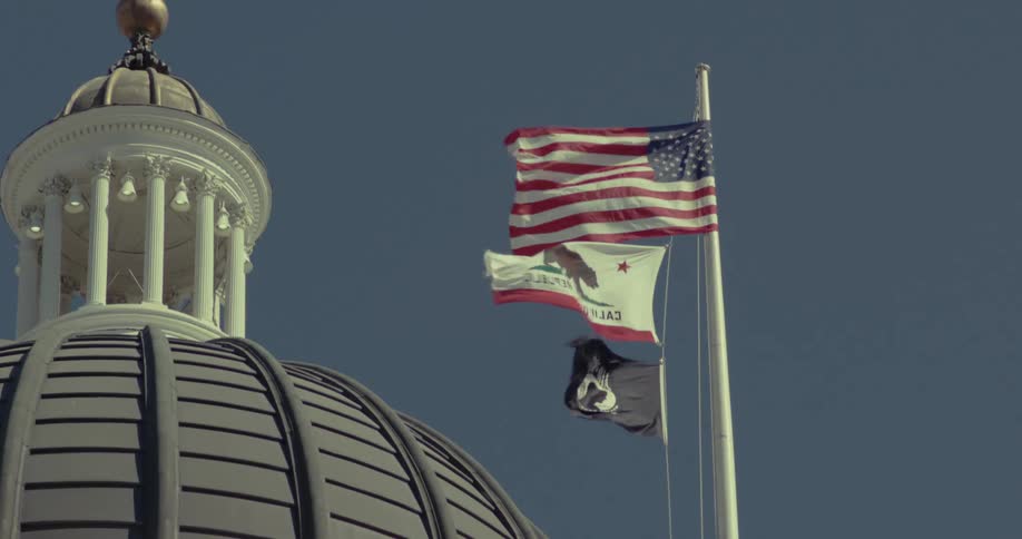 The top of a large gray domed building with a white cupola at its peak set against a clear blue sky with the american flag the california state flag and a dark third flag flying vigorously from a flag pole