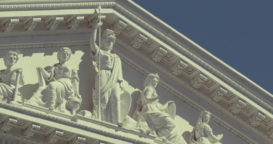 White classical architectural frieze and pediment of a building featuring several statues of robed figures with one standing figure prominent on the left all set sharply against a clear dark blue sky