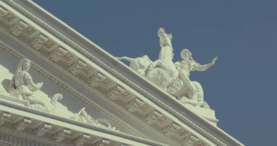 This video shows a static closeup shot of white marble sculptures atop the facade of a classicalstyle building against a clear blue skythe central figures appear to be a group possibly including a person on horseback situated above richly detaile