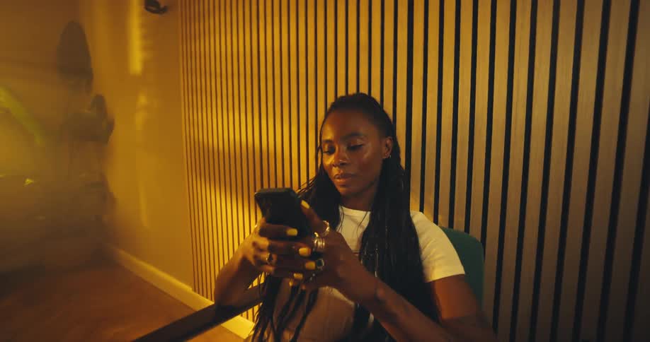This short video shows a young woman sitting in a chair leaning back while intently looking at and scrolling through her mobile phoneshe is dressed casually in a white top and light trousers and the background features striking vertical wooden slats