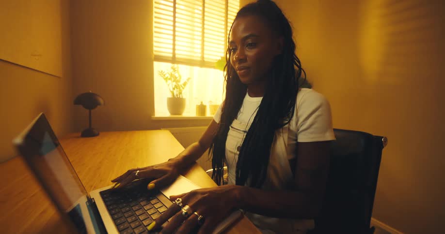 Woman with braided hair typing on laptop at wooden desk lit by natural light