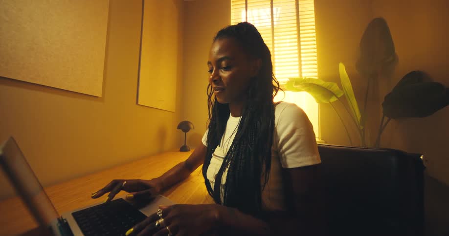 A woman with her long braids pulled back wearing a white shirt is seated at a wooden desk focused on typing on a laptop in a brightly backlit yellowtoned room