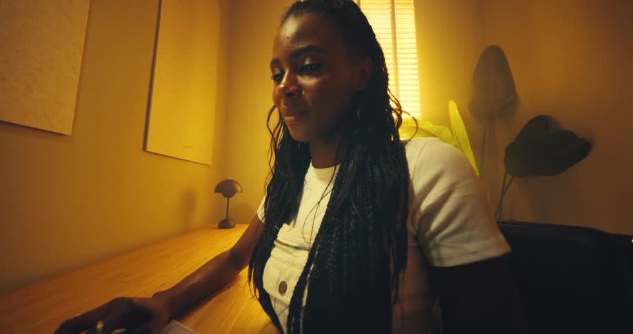 A woman with long braids wearing a white shirt is seated at a wooden desk in a warmly lit yellowwalled room and opens the lid of her laptop to work