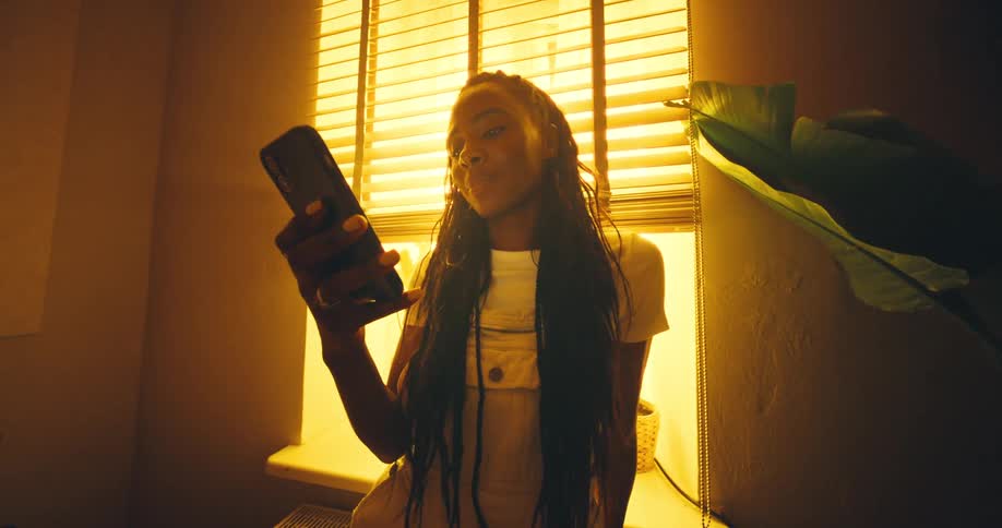 A woman with long braids stands in a strongly backlit warmtoned corner of a room holding her smartphone up and texting
