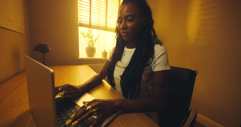This video shows a woman with long braids and bright yellow nail polish sitting at a wooden desk and typing on a laptopshe is in a warmly lit room illuminated by a nearby window with blinds and what appears to be a lamp in the background