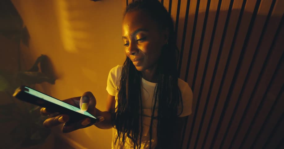 A young woman with braids smiles as she uses her smartphone in a room bathed in warm amber light