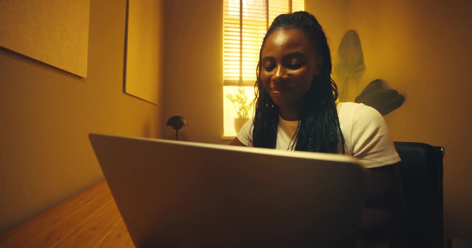 A black woman with long braids is working at a table opens a laptop and looks at the laptop smiling while golden light is streaming in through blinds visible in the background
