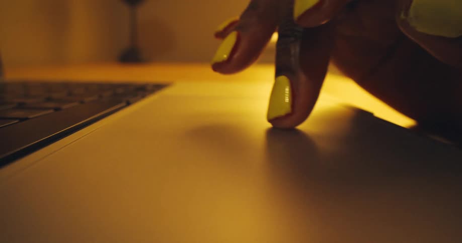 A darkskinned hand with bright yellow nail polish using the trackpad of a laptop illuminated by warm golden lighting that contrasts with the dark keyboard keys visible in the blurred foreground