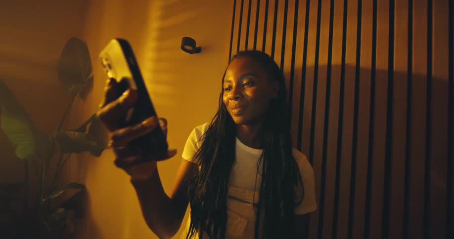 A smiling young woman holds up her smartphone in a room filled with warm golden light